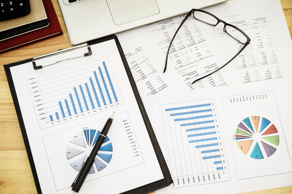 Close-up of financial and business reports with bar charts and pie graphs displayed on a clipboard and papers. A pen and a pair of glasses are placed on top of the documents, along with a laptop in the background. The image highlights data analysis, business performance review, and financial planning.